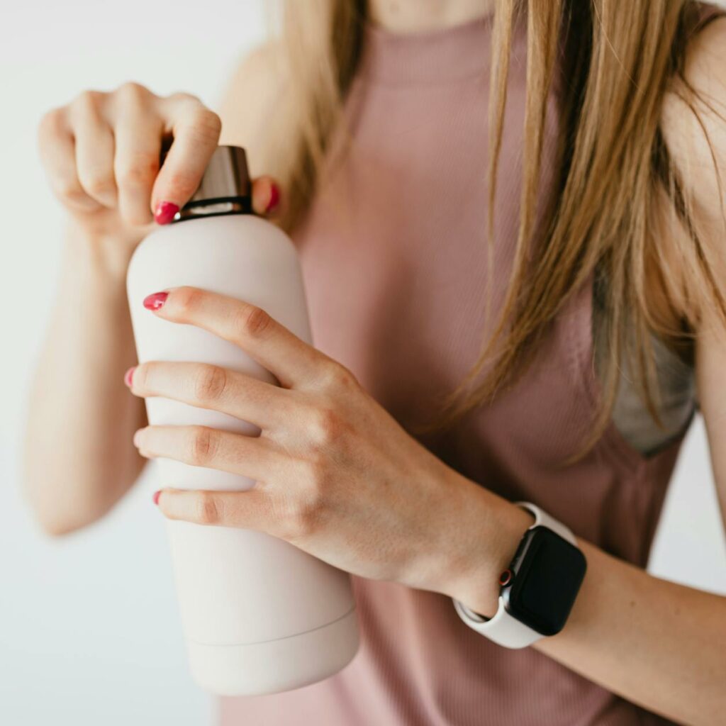 Crop faceless young female in casual outfit wearing smart watch opening cosmetic bottle on white background