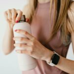 Crop faceless young female in casual outfit wearing smart watch opening cosmetic bottle on white background