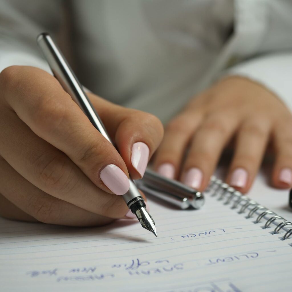 Woman in White Long Sleeved Shirt Holding a Pen Writing on a Paper