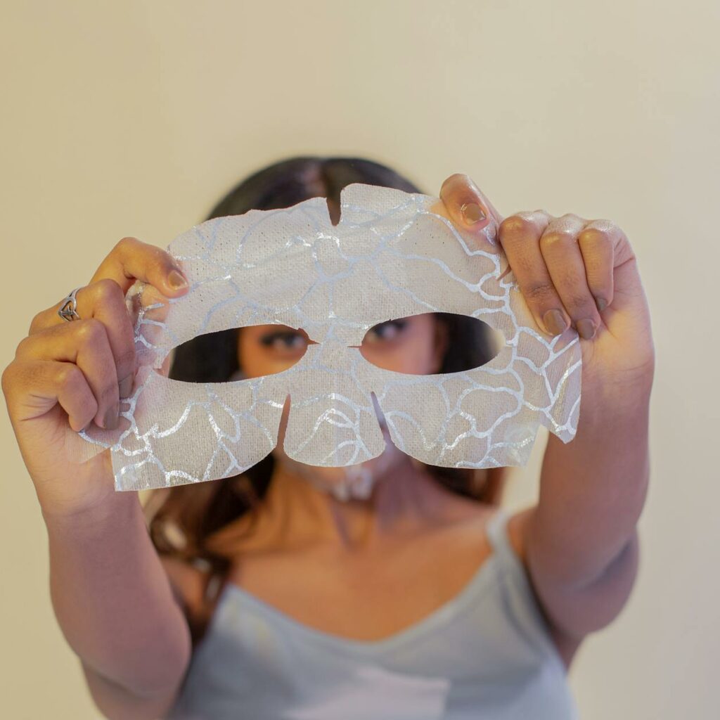 Calm ethnic female showing rejuvenating eye mask sheet in outstretched arms and looking at camera in studio