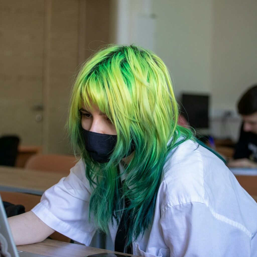 A girl with green hair and a face mask is using a laptop