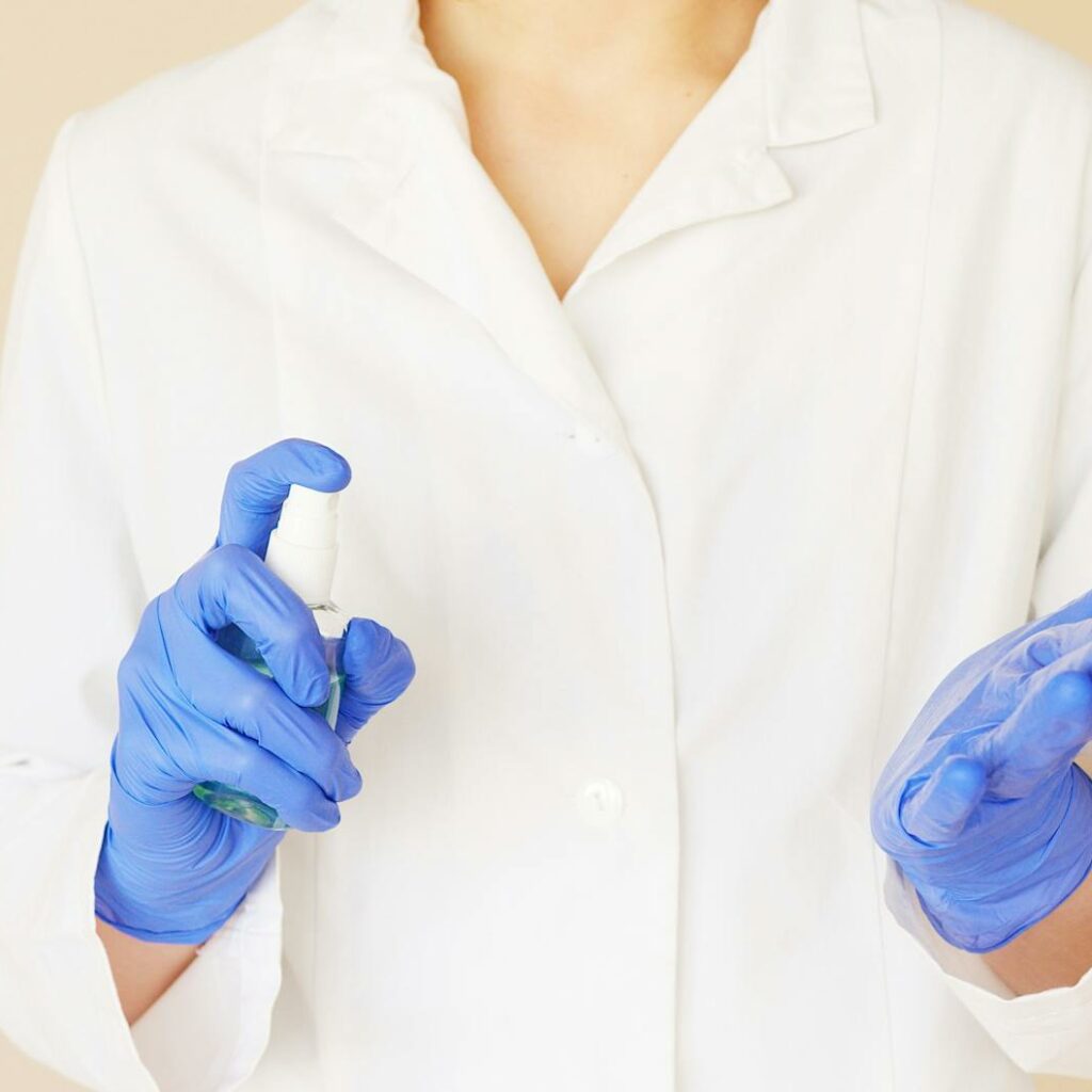 Crop unrecognizable hospital worker in white uniform spraying antiseptic liquid over hands in blue latex gloves