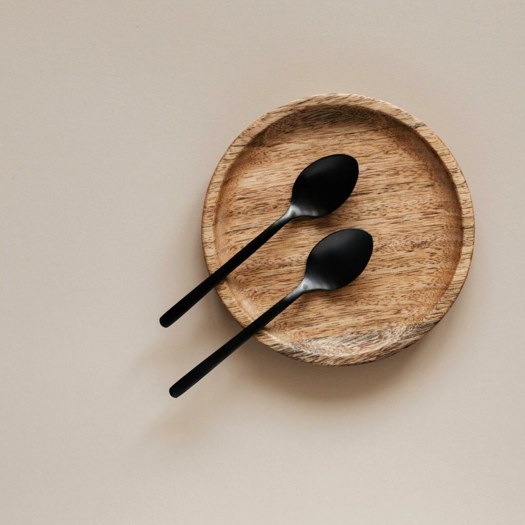 Top view of composition of round wooden plate with smooth surface and similar black metal spoons on beige surface