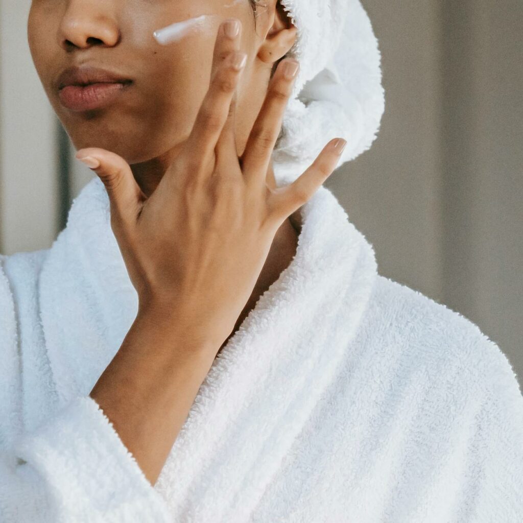 Crop anonymous female in white bathrobe with towel on head applying facial moisturizing cream on face while standing in bathroom